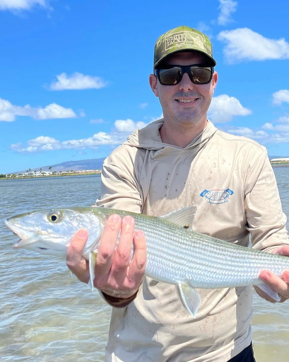Man holding a bonefish by a body of water with a clear sky