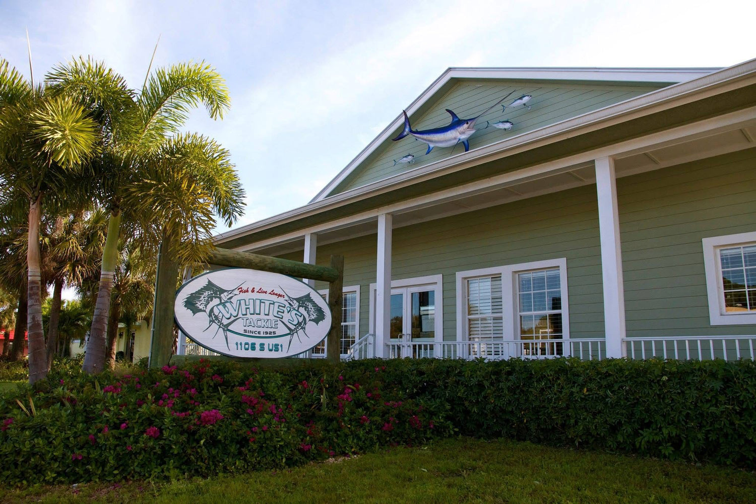 White's Tackle store front Green building with a sign and decorative fish on the roof, surrounded by palm trees and flowers.