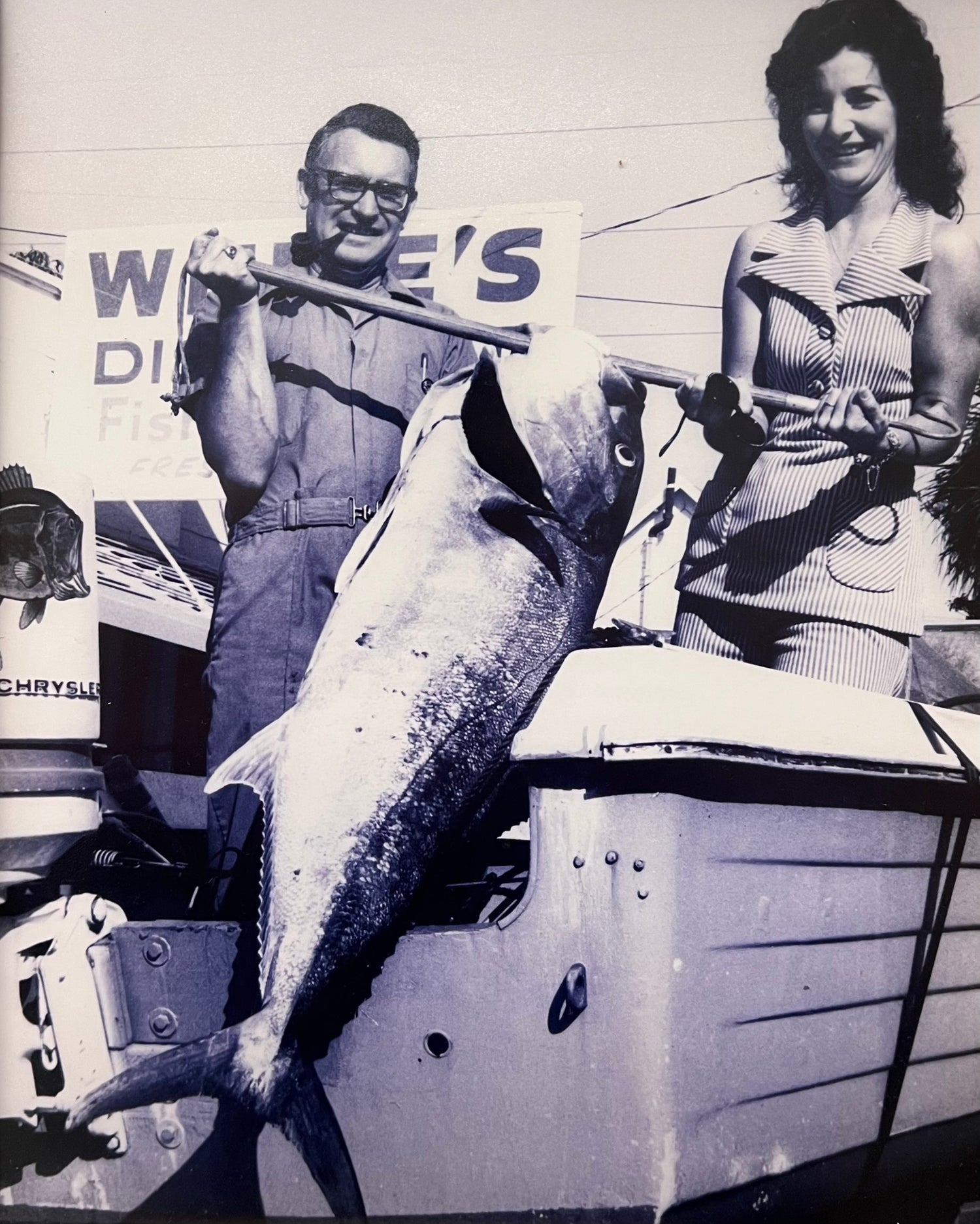Black and white photo of a man and woman holding a large fish, with a boat and 'White's' sign in the background.