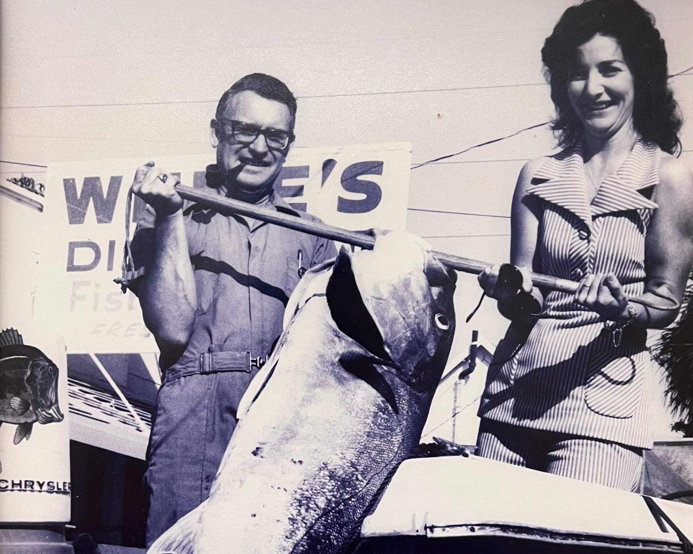 Black and white photo of a man and woman holding a large fish, with a boat and 'White's' sign in the background.