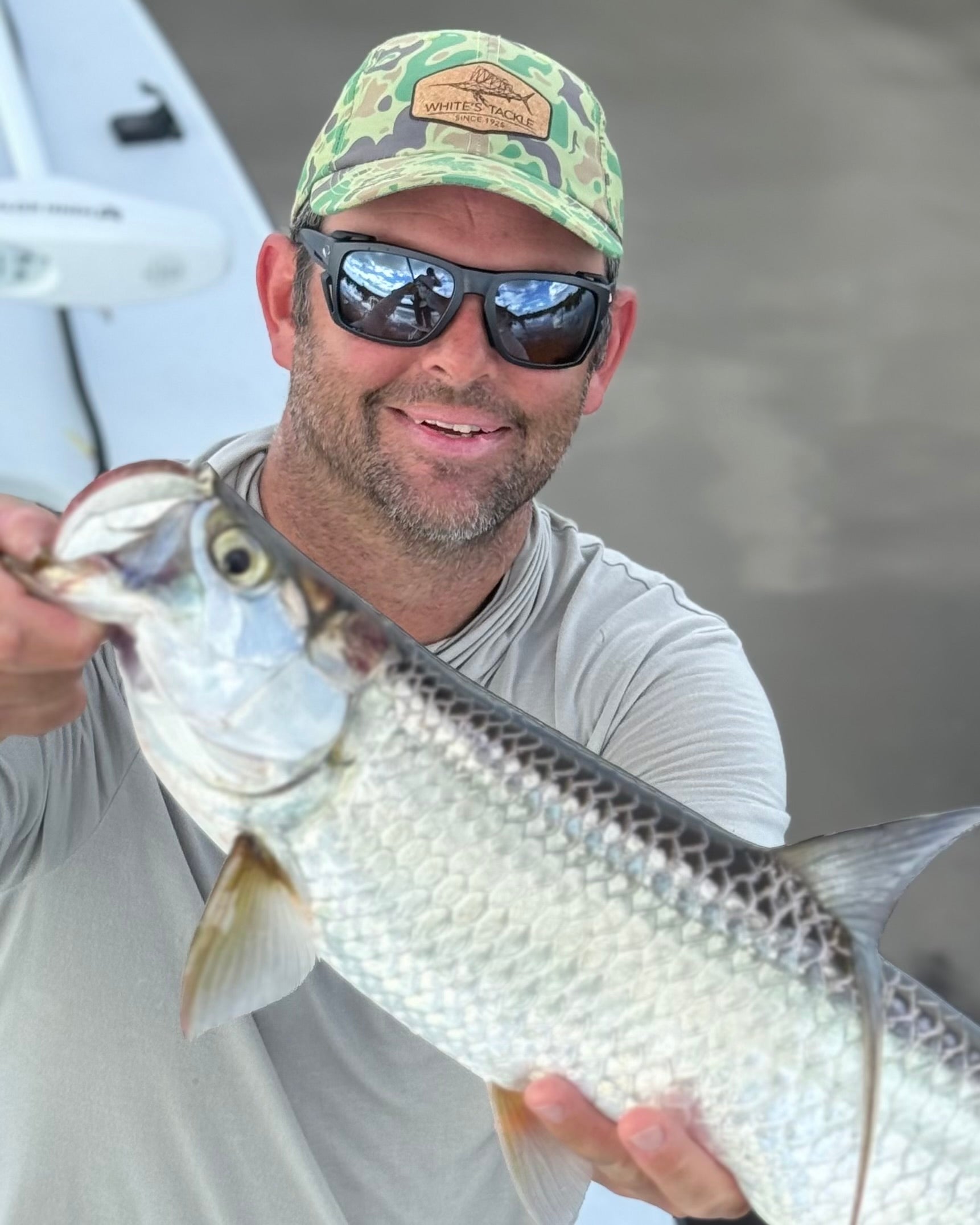 Man holding a small tarpon, wearing sunglasses and a cap, with a blurred background